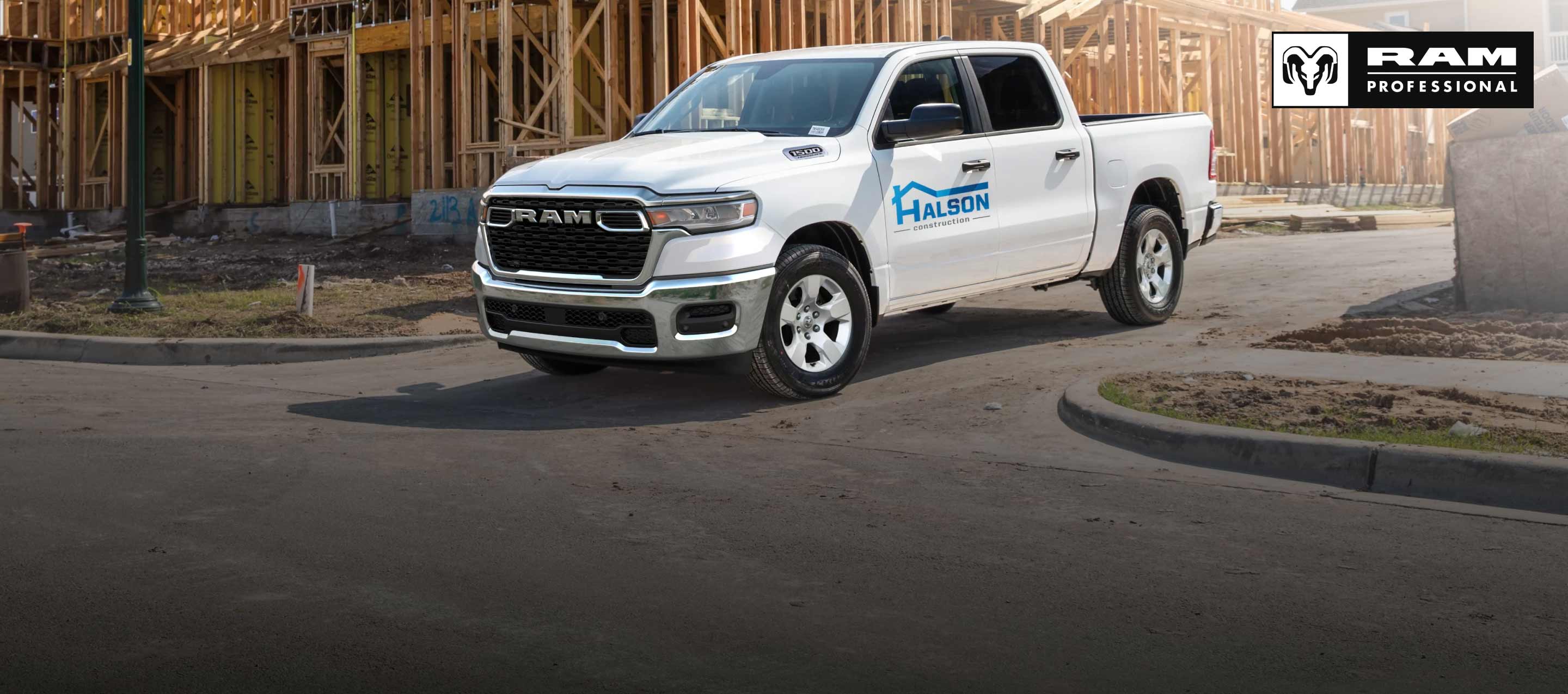 Una Ram 1500 Tradesman 4x4 Crew Cab 2026 blanca, con gráfico de una compañía de construcción en la puerta del conductor, saliendo de una obra de construcción comercial. Ram Professional.