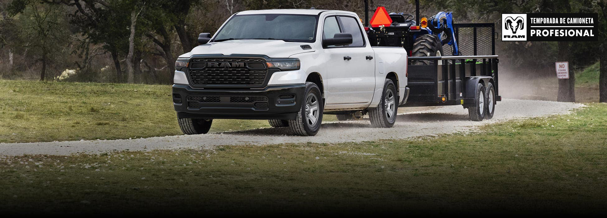 Una Ram 1500 Tradesman 4x4 Crew Cab 2026 blanca remolcando un tractor en un remolque para paisajismo mientras circula por una carretera de tierra. Temporada de camionetas Ram Professional.