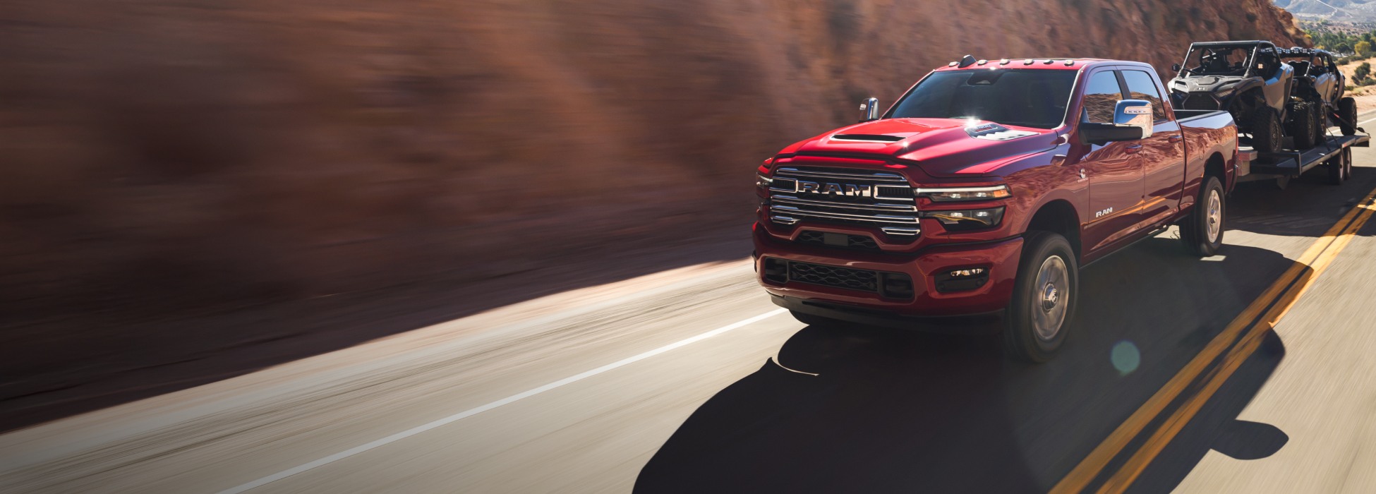 Una Ram 2500 Laramie 4x4 Crew Cab 2026 roja circulando por una carretera en las montañas, mientras remolca dos ATV.
