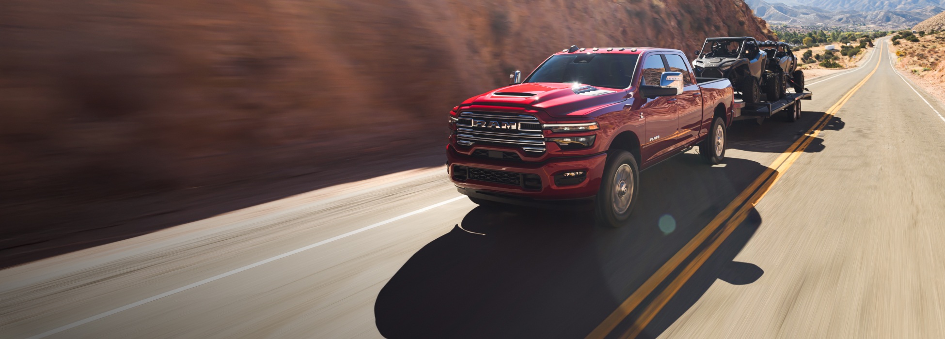 Una Ram 2500 Laramie 4x4 Crew Cab 2026 roja circulando por una carretera en las montañas, mientras remolca dos ATV.