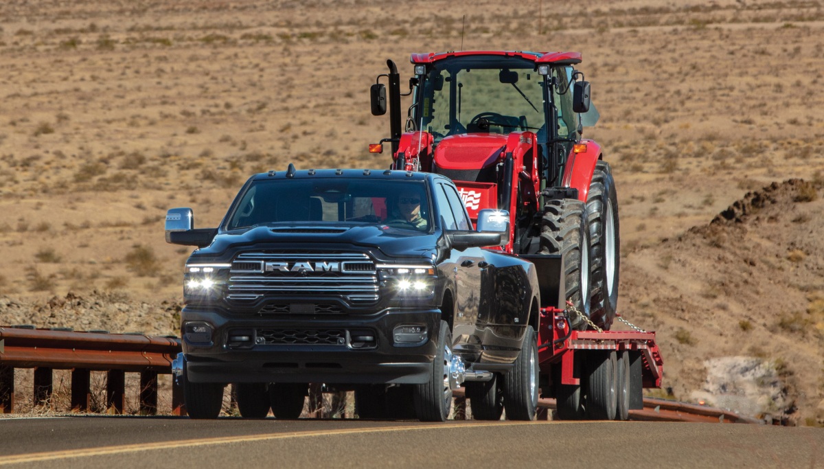 Se muestra una Ram 3500 Limited Longhorn 4x4 Crew Cab 2026 negra acarreando un vehículo de construcción en un remolque de plataforma plana largo, circulando por un camino de montaña.