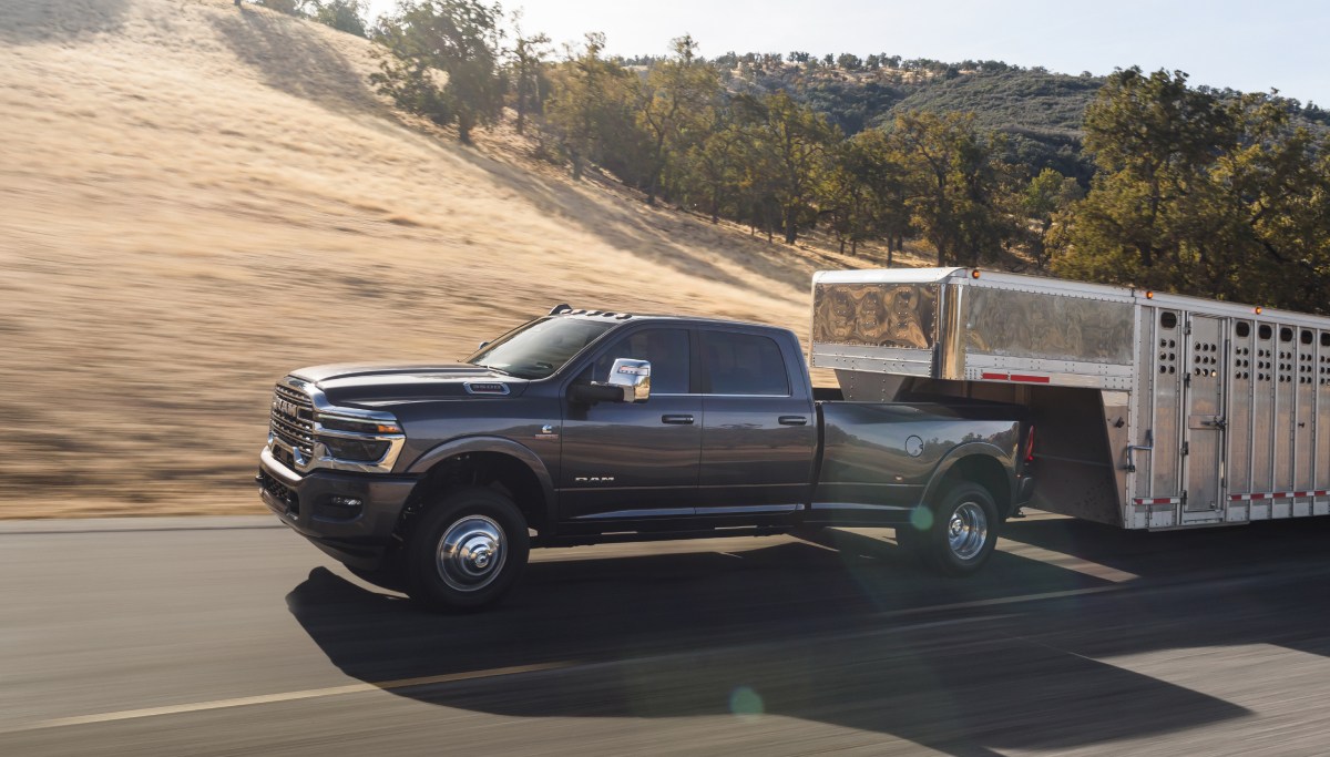 Se muestra una Ram 3500 Limited Longhorn 4x4 Crew Cab 2026 gris acarreando un remolque de quinta rueda grande para ganado mientras circula por una carretera.