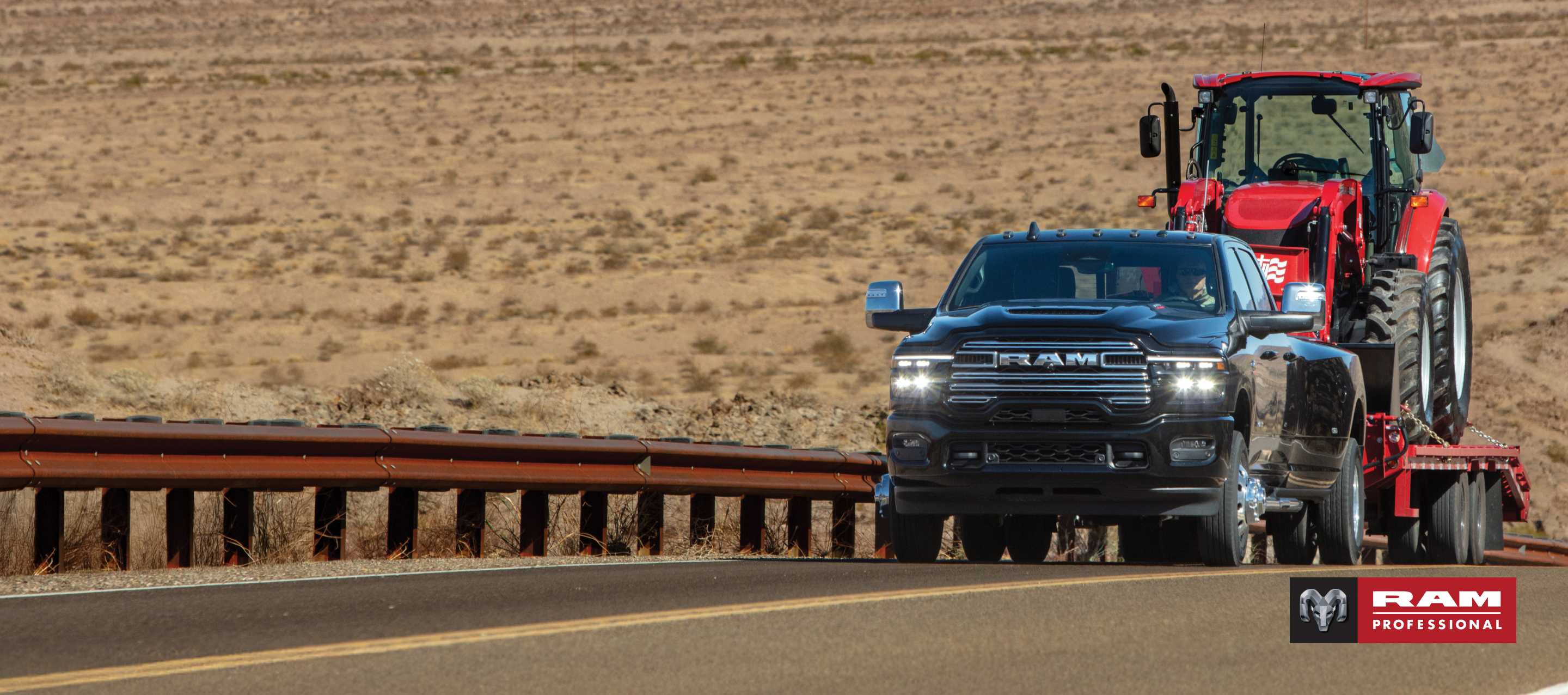 Se muestra una Ram 3500 Limited Longhorn 4x4 Crew Cab 2026 negra acarreando un vehículo de construcción en un remolque de plataforma plana largo, circulando por un camino de montaña. Ram Professional
