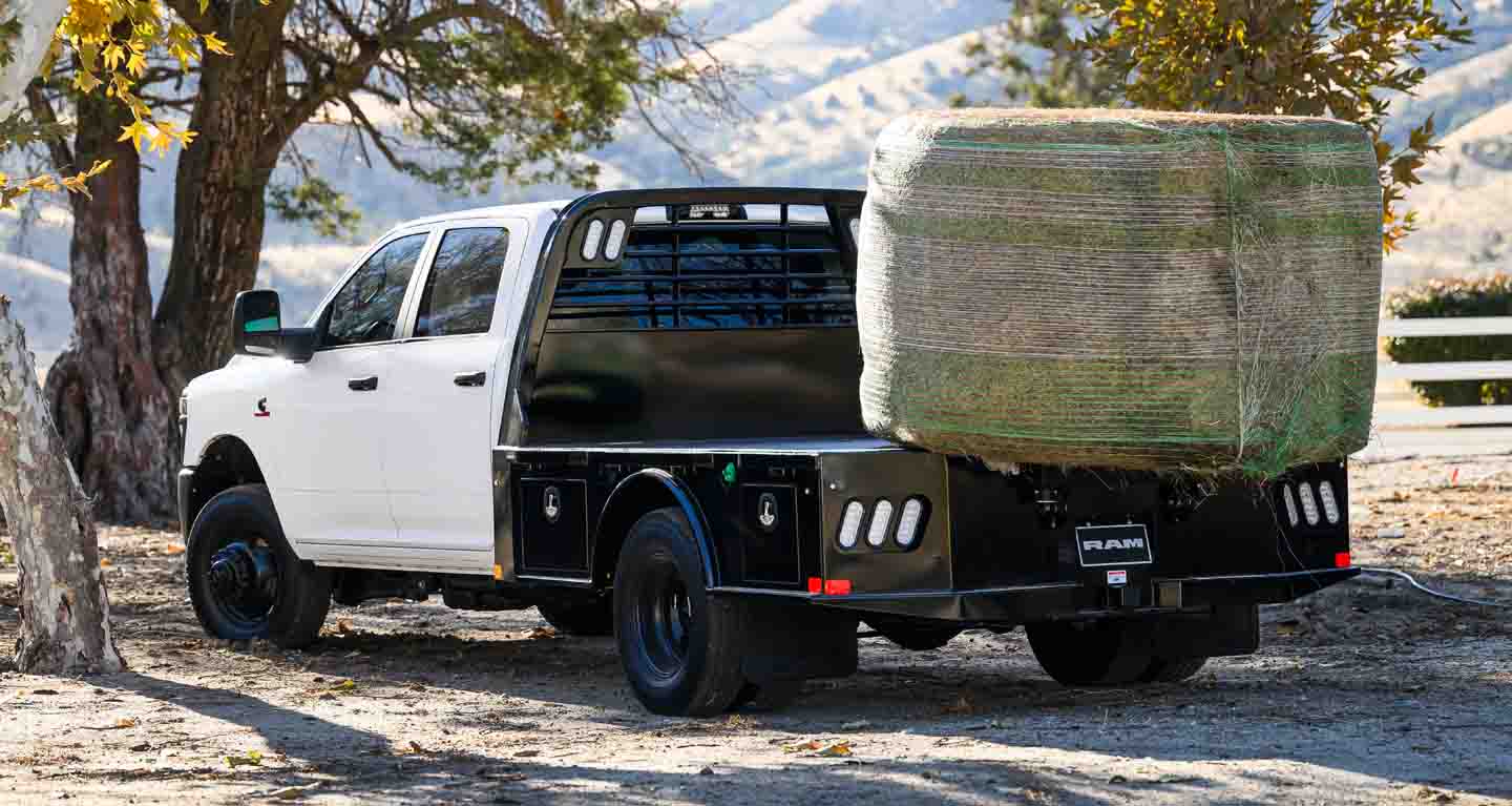 Se muestra Una Ram 3500 Tradesman Chassis Cab Crew Cab 2026 blanca, con un gran fardo de heno sobre su caja tipo plataforma, estacionada en una granja.