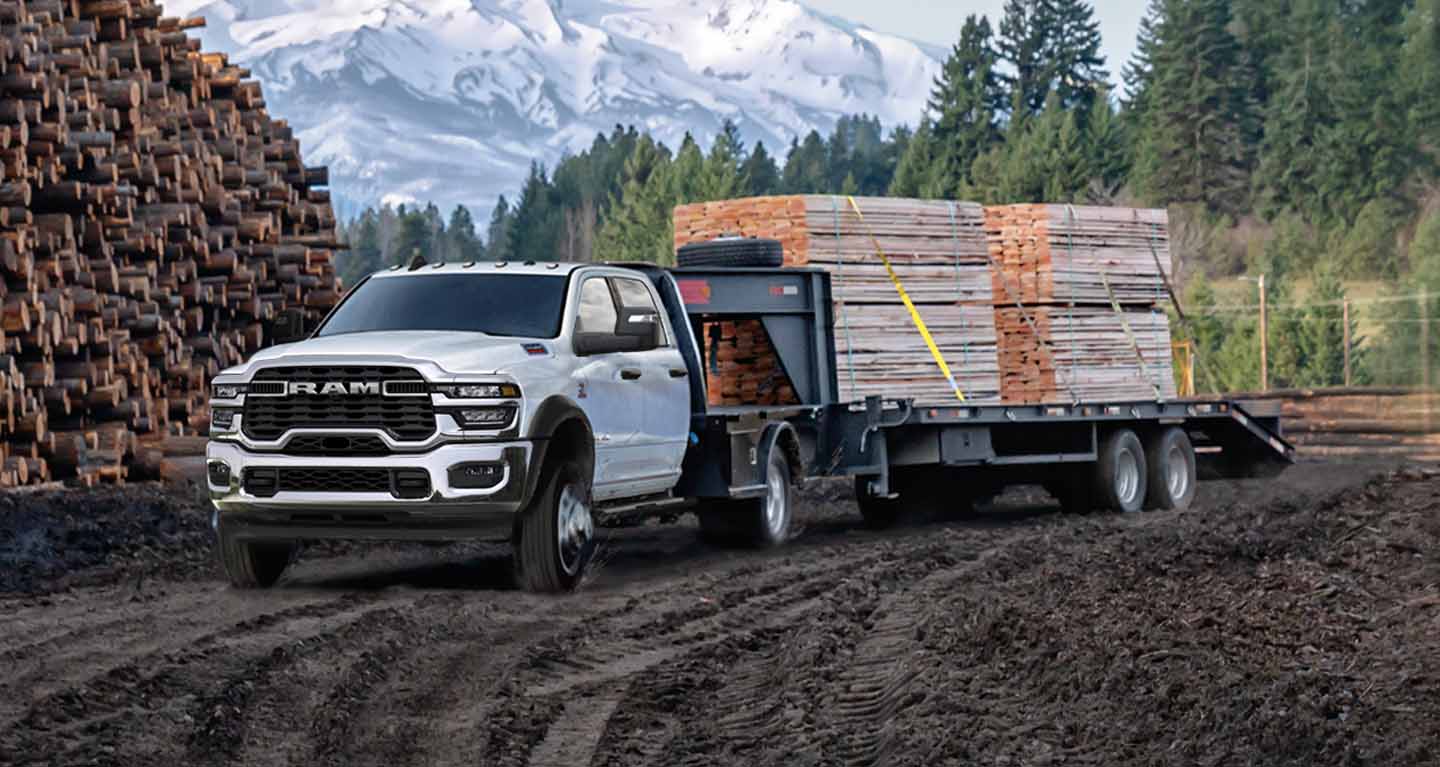 Se muestra Una Ram 5500 Tradesman Chassis Cab Crew Cab 2026 blanca, con un remolque cuello de cisne lleno de madera, estacionada junto a un bosque y un gran montón de troncos cortados.
