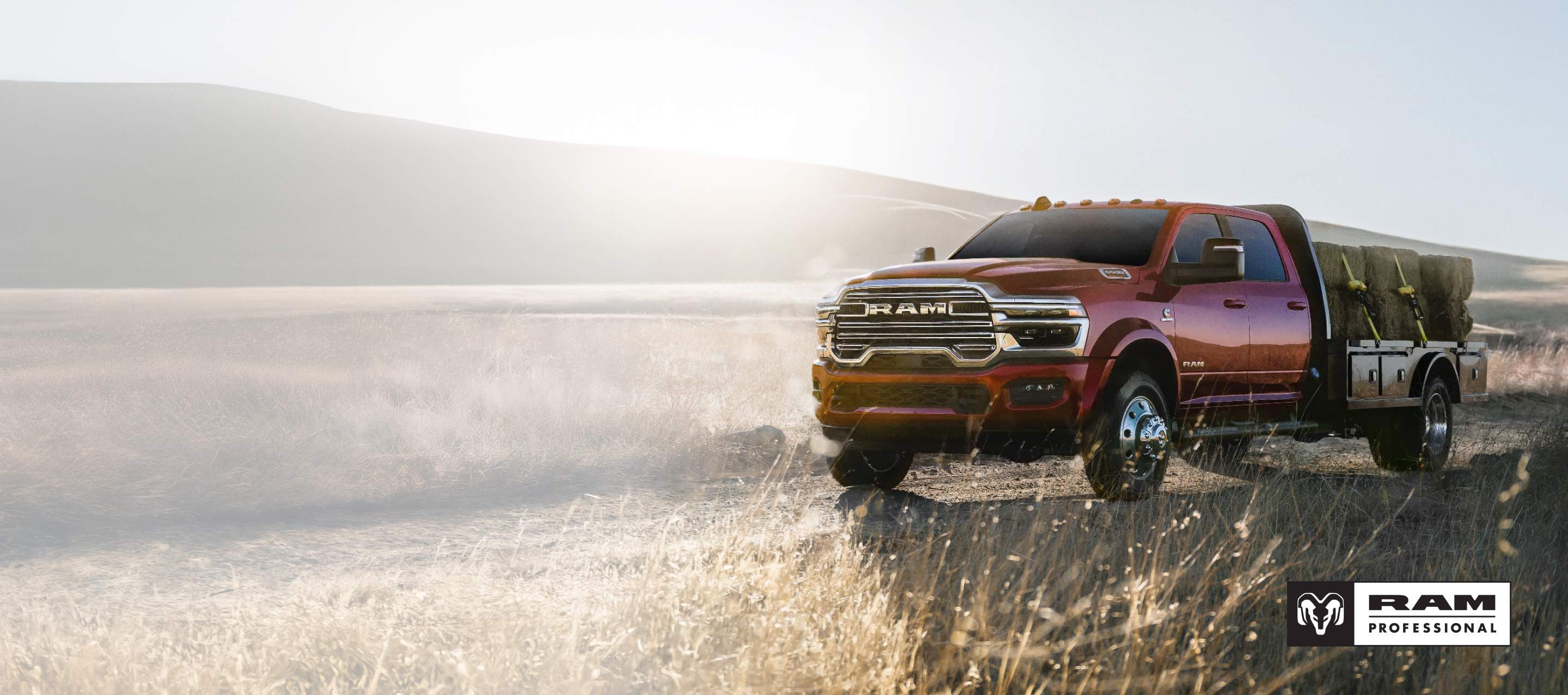Una Ram 5500 Laramie Chassis Cab Crew Cab 2026 roja trasladando varios fardos de heno grandes en una mejora de plataforma larga mientras circula por un camino de tierra de una granja. Ram Professional.
