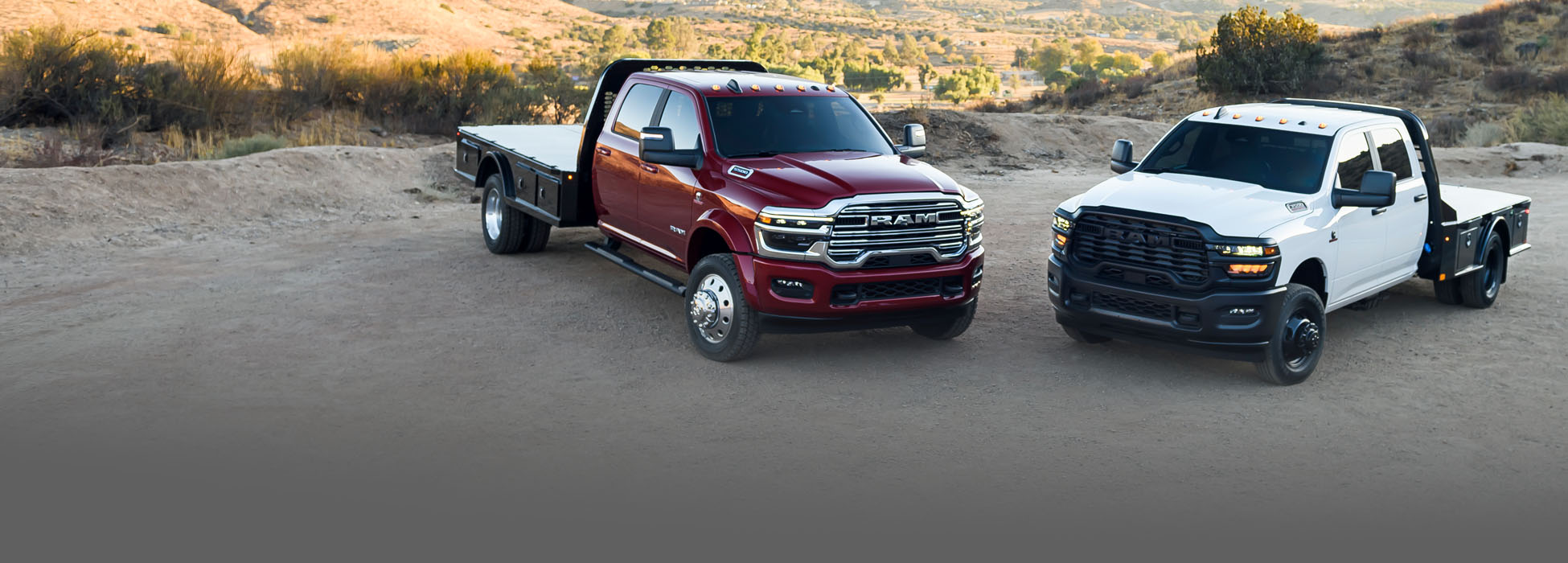 Dos Ram Chassis Cab, ambas con cajas tipo plataforma, estacionadas en un claro. A la izquierda, una Ram 5500 Laramie Chassis Cab Crew Cab 2026 roja y, a la derecha, una Ram 3500 Tradesman Chassis Cab Crew Cab 2026 blanca.