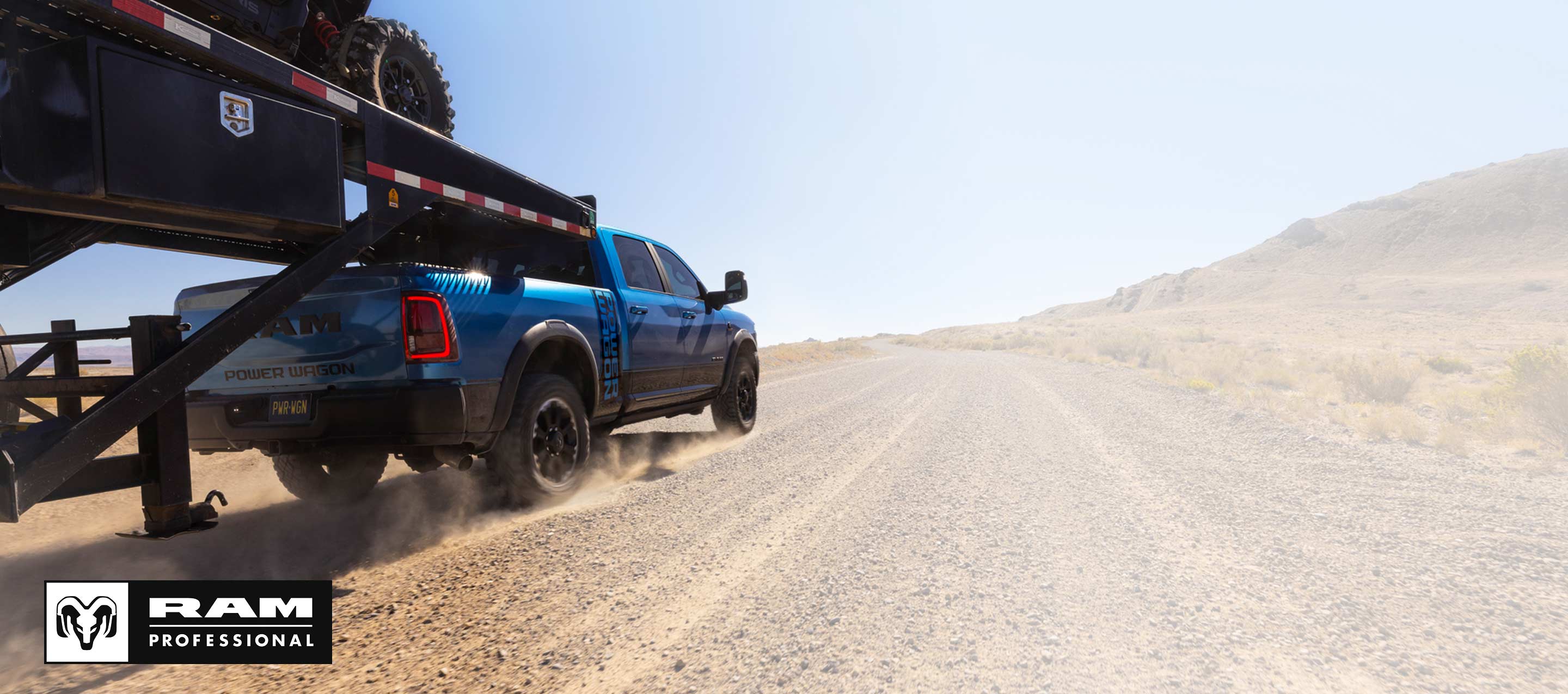 Una Ram 2500 Power Wagon 4x4 Crew Cab 2026 azul para trabajo pesado acarreando un ATV en un remolque de quinta rueda y plataforma plana mientras circula por un camino de tierra. Ram Professional.