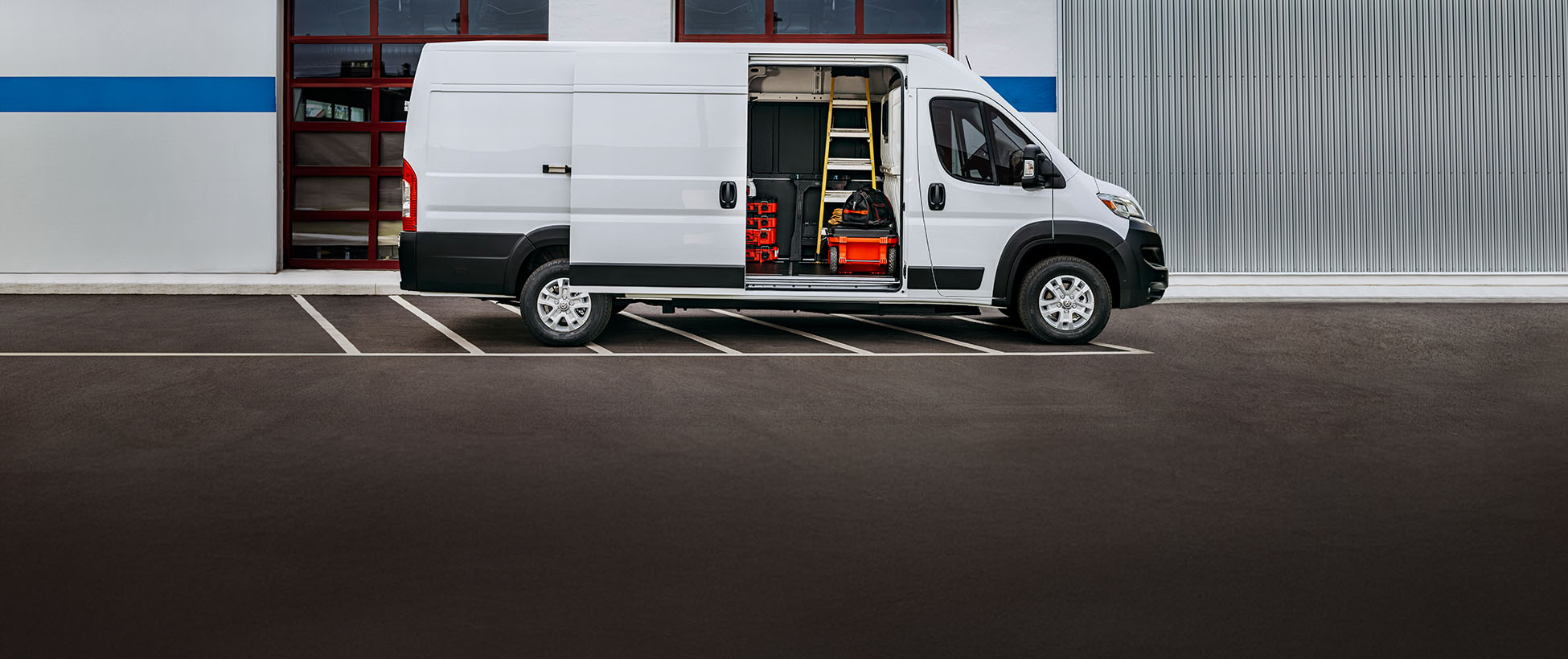 Perfil del lado del pasajero de una Ram ProMaster EV 3500 SLT High Roof Cargo Van 2026 blanca estacionada al lado de un garaje comercial con la puerta lateral corrediza abierta, lo que permite ver herramientas, equipos y una escalera en el interior.