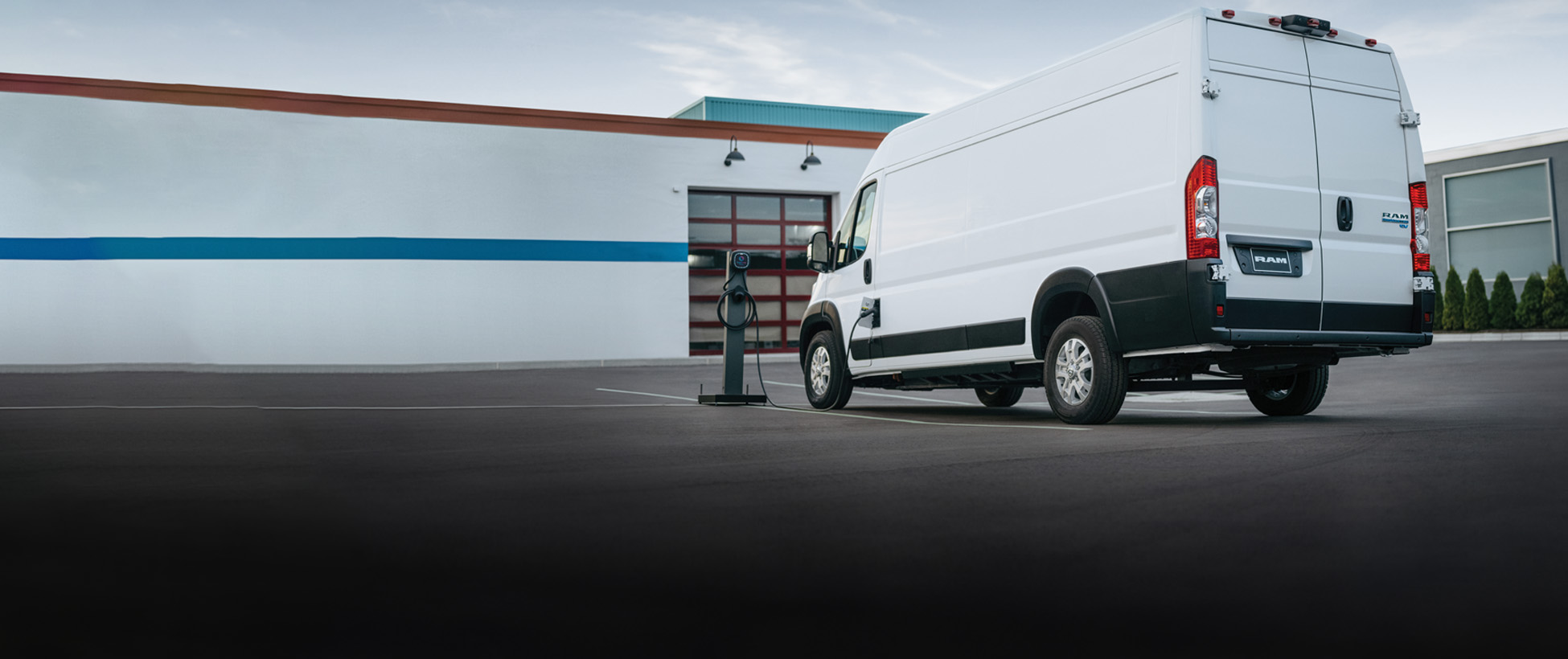 Una Ram ProMaster EV 3500 SLT High Roof Cargo Van 2026 blanca estacionada al lado de una estación de carga pública con el cable de carga enchufado en el puerto de carga de la van.