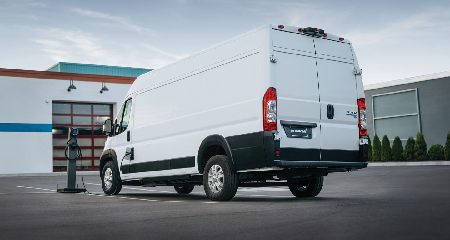 Se muestra una Ram ProMaster EV 3500 SLT High Roof Cargo Van 2026 blanca estacionada al lado de una estación de carga pública con el cable de carga enchufado en el puerto de carga de la van.