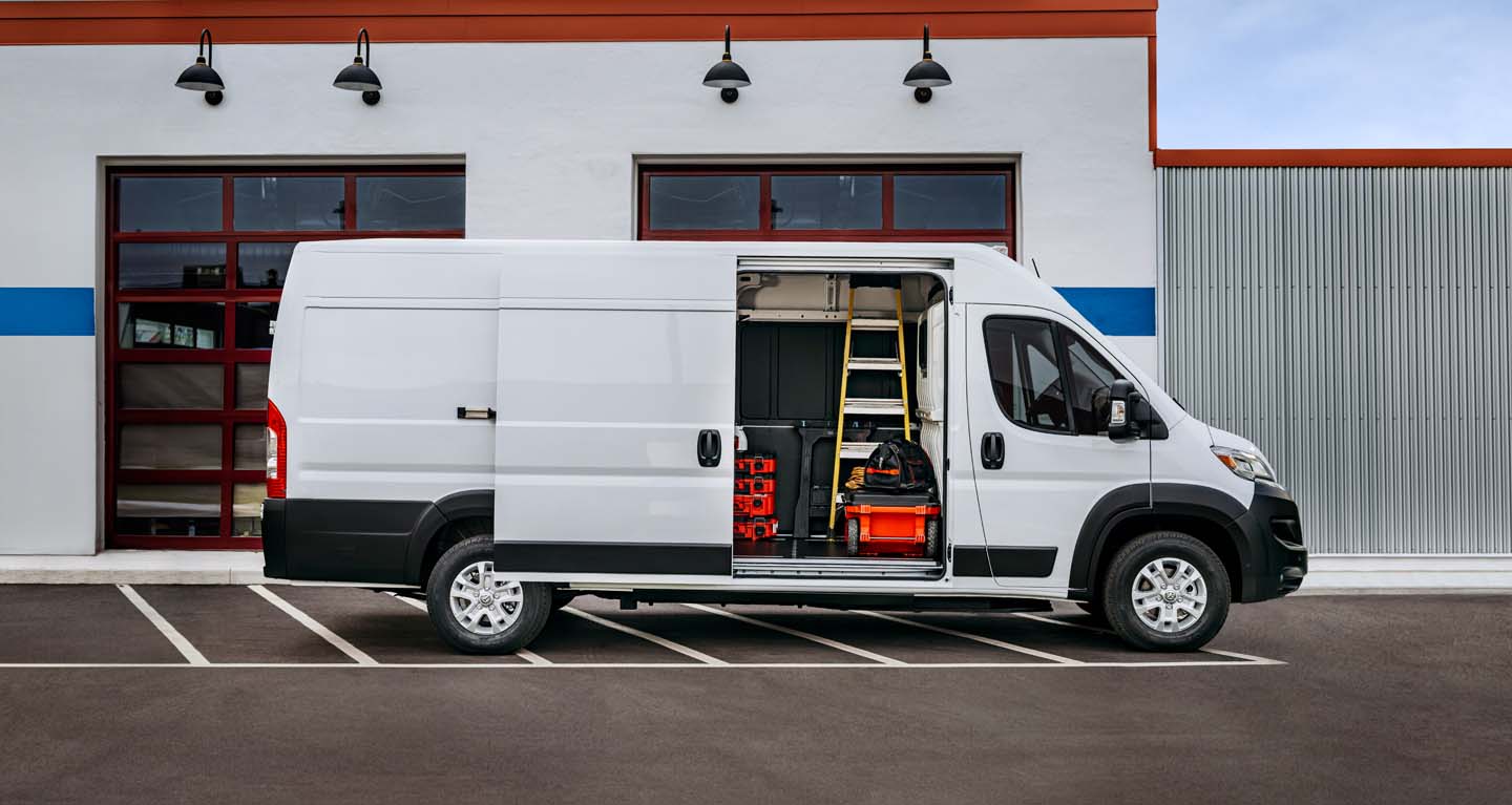 Se muestra un perfil del lado del pasajero de una Ram ProMaster EV 3500 SLT High Roof Cargo Van 2026 blanca estacionada al lado de un garaje comercial con la puerta lateral corrediza abierta, lo que permite ver herramientas, equipos y una escalera en el interior.
