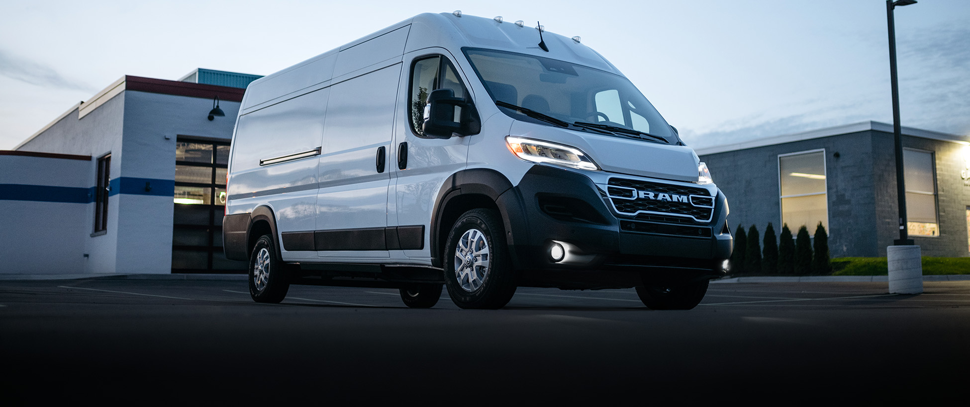 Ángulo delantero del lado del pasajero de una Ram ProMaster EV 3500 SLT High Roof Cargo Van 2026 blanca con los faros delanteros y antiniebla encendidos, estacionada cerca de un garaje comercial al atardecer.