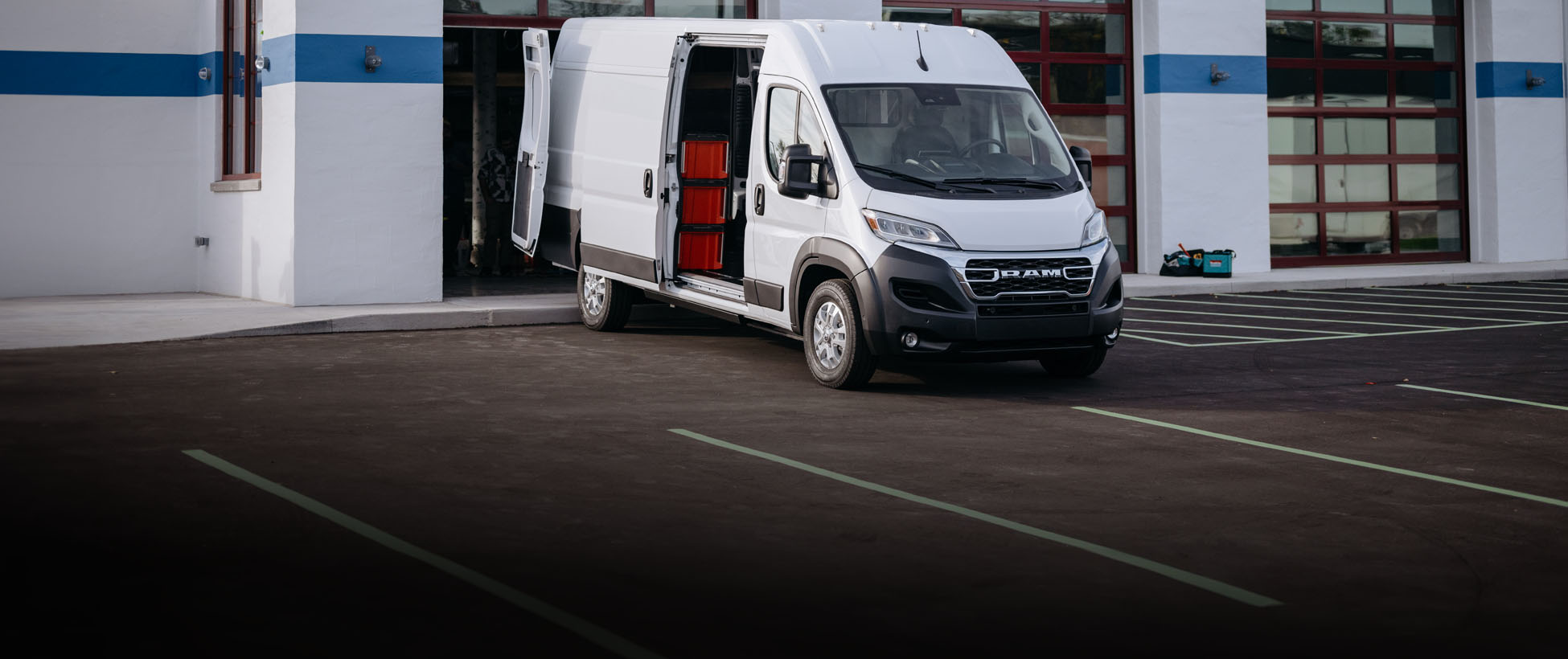 Ángulo delantero del lado del pasajero de una Ram ProMaster EV 3500 SLT High Roof Cargo Van 2026 blanca con las puertas lateral y trasera abiertas, estacionada cerca de la puerta de un garaje abierta de un edificio comercial.
