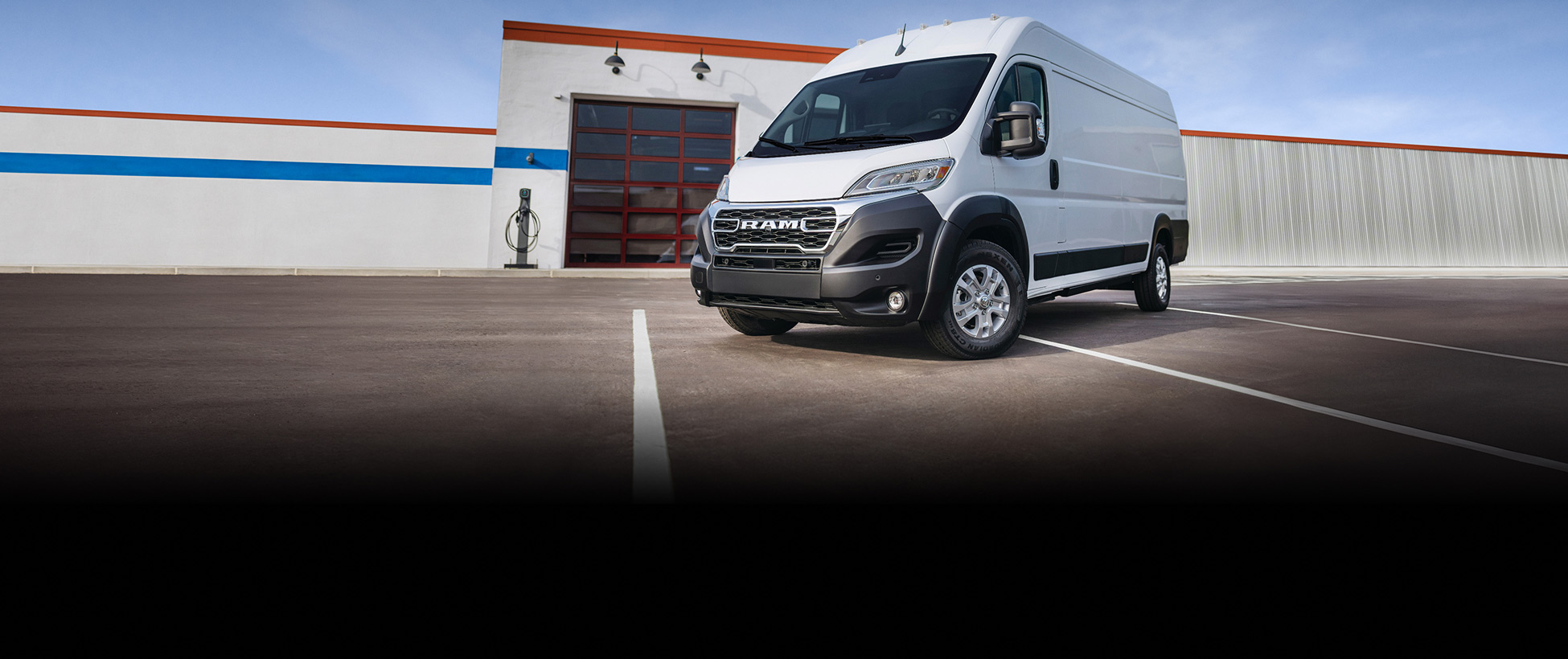 Ángulo delantero del lado del conductor de una Ram ProMaster EV 3500 SLT Plus High Roof Cargo Van 2026 blanca estacionada cerca de un garaje comercial.