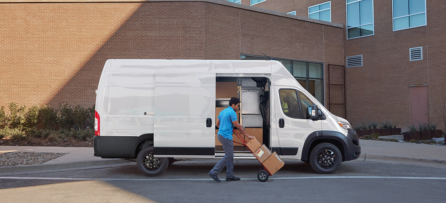 Se muestra un hombre empujando paquetes en una carretilla cerca de la puerta lateral corrediza abierta de una Ram ProMaster 3500 SLT Plus Cargo Van Super High Roof 2026 blanca con el paquete de apariencia negro.