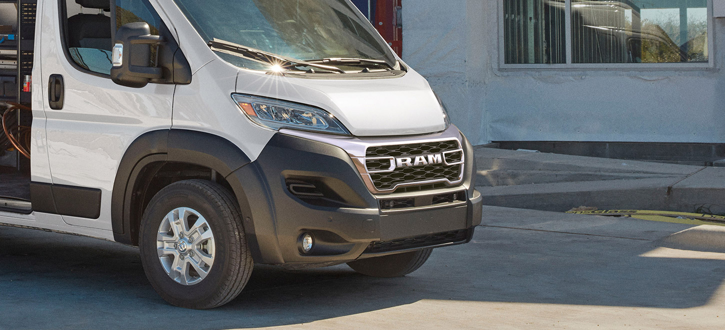 Se muestra el ángulo delantero del lado del pasajero de una Ram ProMaster 2500 SLT Cargo Van High Roof 2026 blanca estacionada al lado de un edificio comercial.