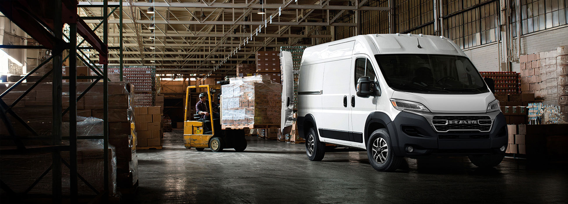 Un ángulo delantero del lado del pasajero de una Ram ProMaster 2500 SLT Cargo Van High Roof 2026 blanca estacionada en un depósito con las puertas traseras totalmente abiertas y un montacargas con una pila de cajas circulando hacia la parte trasera de la van.