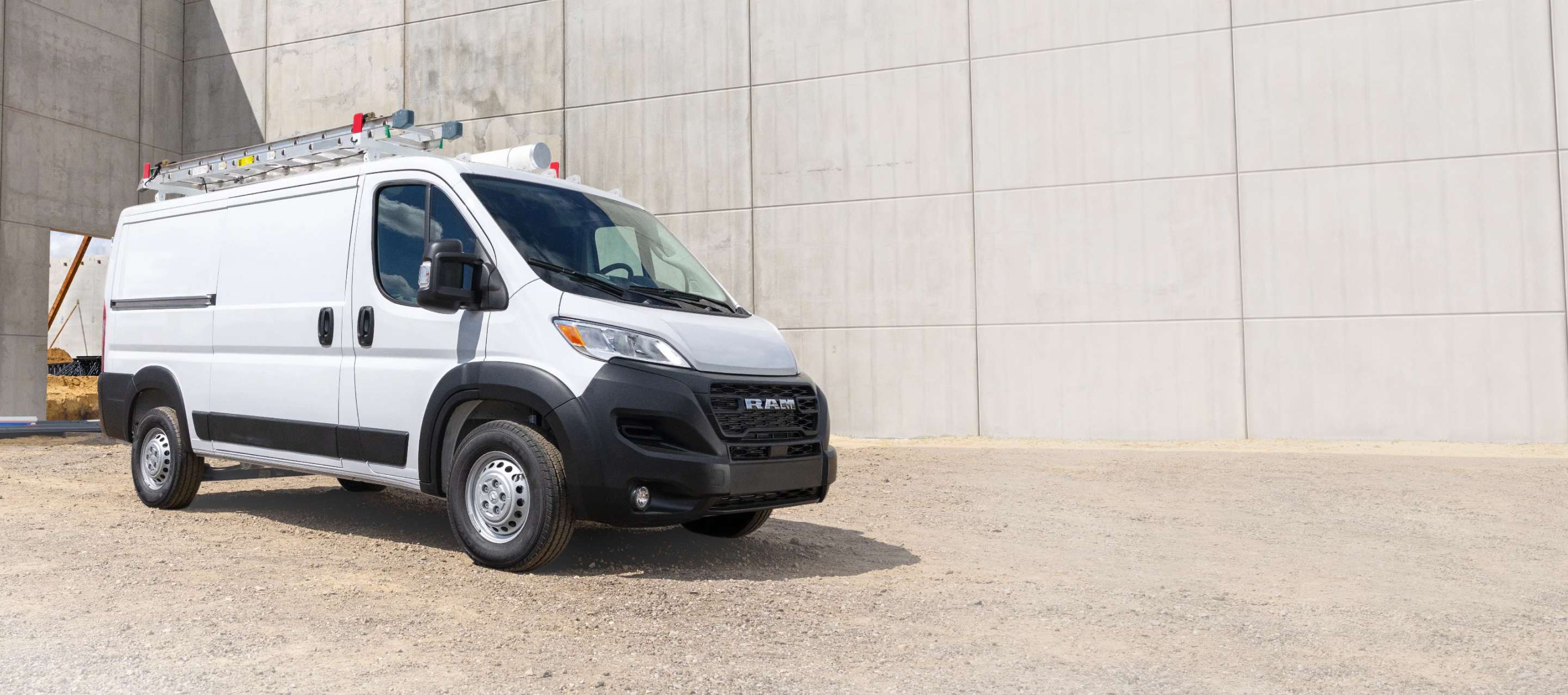 Una Ram ProMaster Tradesman Standard Roof Cargo Van 2026 blanca con una escalera montada en el portaequipajes del techo, en un sitio de construcción, fotografiada de perfil en ángulo.