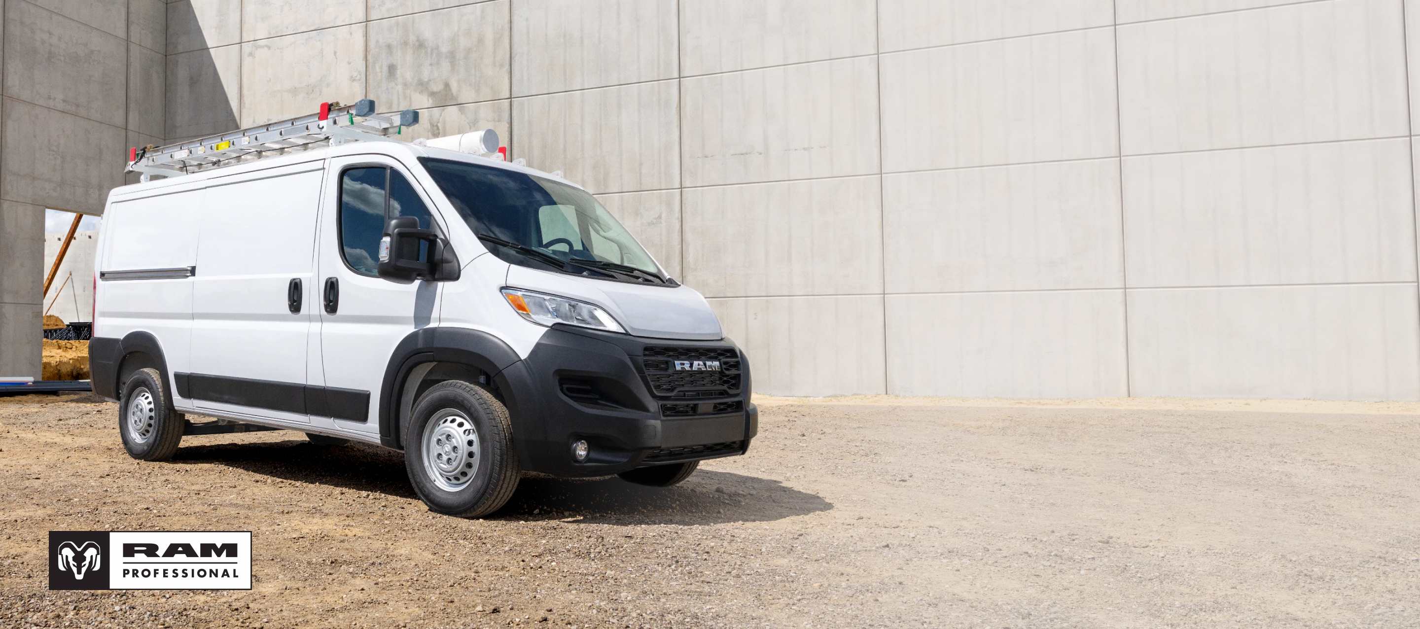 Una Ram ProMaster Tradesman Standard Roof Cargo Van 2026 blanca con una escalera montada en el portaequipajes del techo, en un sitio de construcción, fotografiada de perfil en ángulo. Ram Professional.