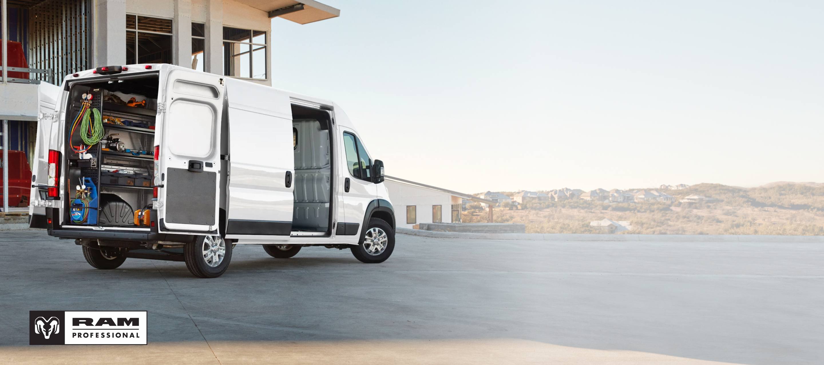 La parte trasera de una Ram ProMaster 3500 SLT High Roof Cargo Van 2026 blanca con la puerta deslizable del lado del pasajero y las puertas traseras abiertas por las cuales se observan estantes llenos de herramientas. Ram Professional.