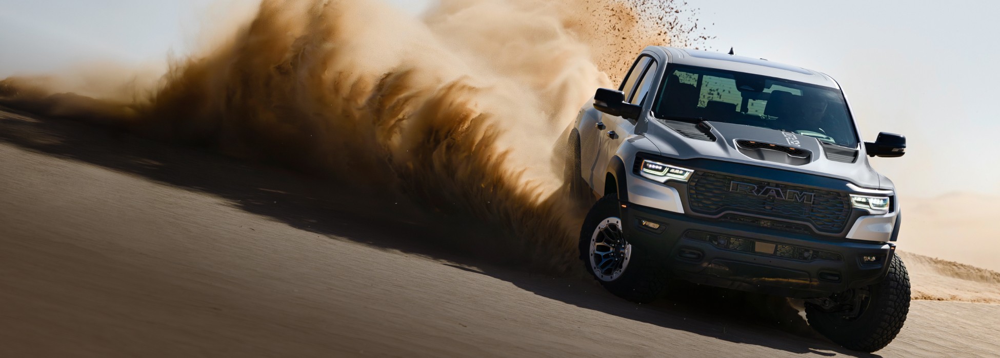 Ángulo frontal de una Ram 1500 RHO 4x4 Crew Cab 2026 plateada conduciendo fuera de carretera en una colina del desierto, con polvo y arena levantándose de sus neumáticos.