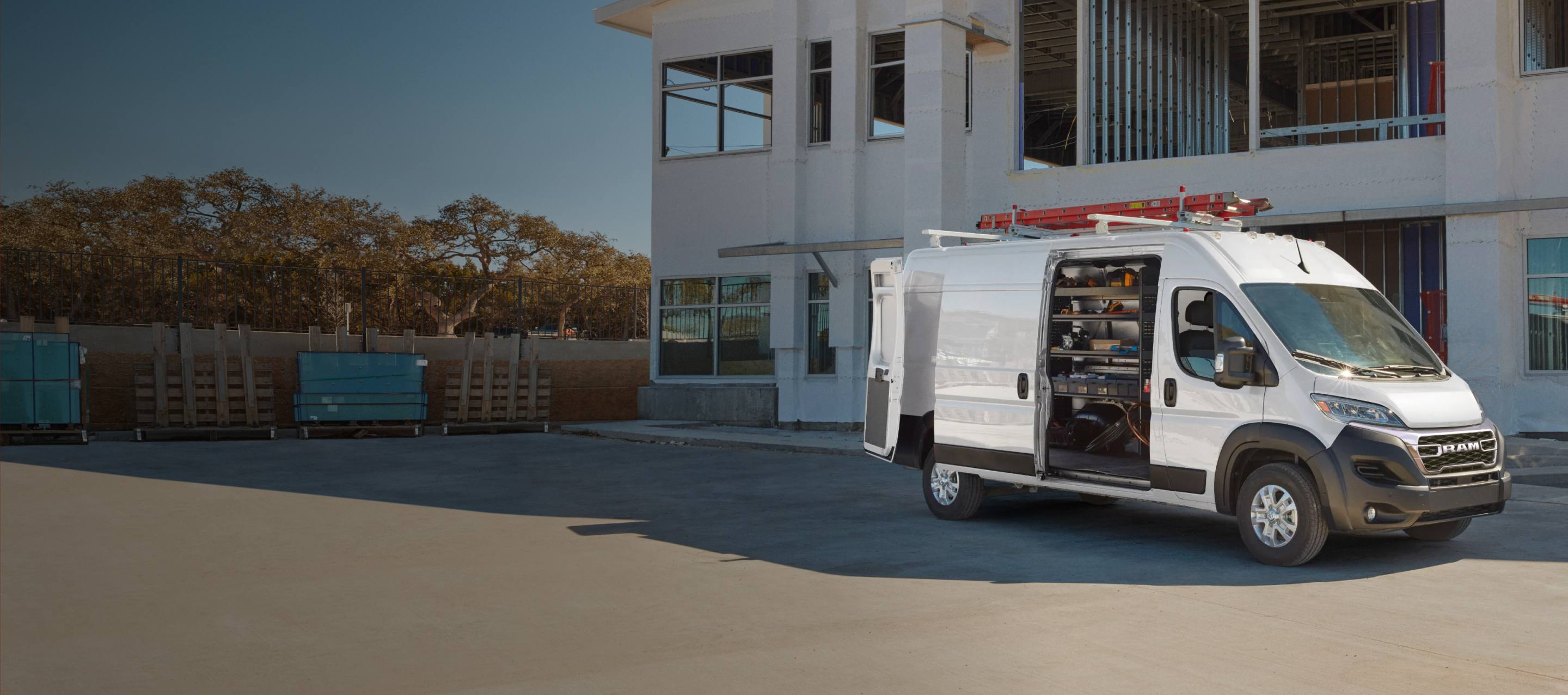 Una Ram ProMaster 3500 SLT High Roof Cargo Van 2027 blanca, estacionada en el exterior de un edificio comercial con una escalera roja en la parrilla portaequipaje y la puerta corrediza del lado del pasajero abierta, mostrando estantes personalizados llenos de equipos. Diseñada para profesionales. Ram Professional.