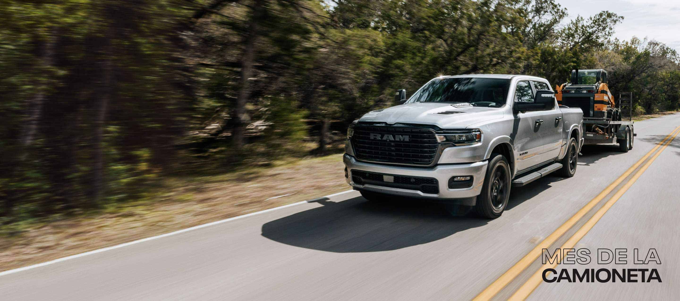 Una Ram 1500 Laramie 4x4 Crew Cab 2025 plateada circulando por una carretera rural, acarreando equipo para movimiento de tierras en un remolque basculante. Evento de ventas Mes de las camionetas.