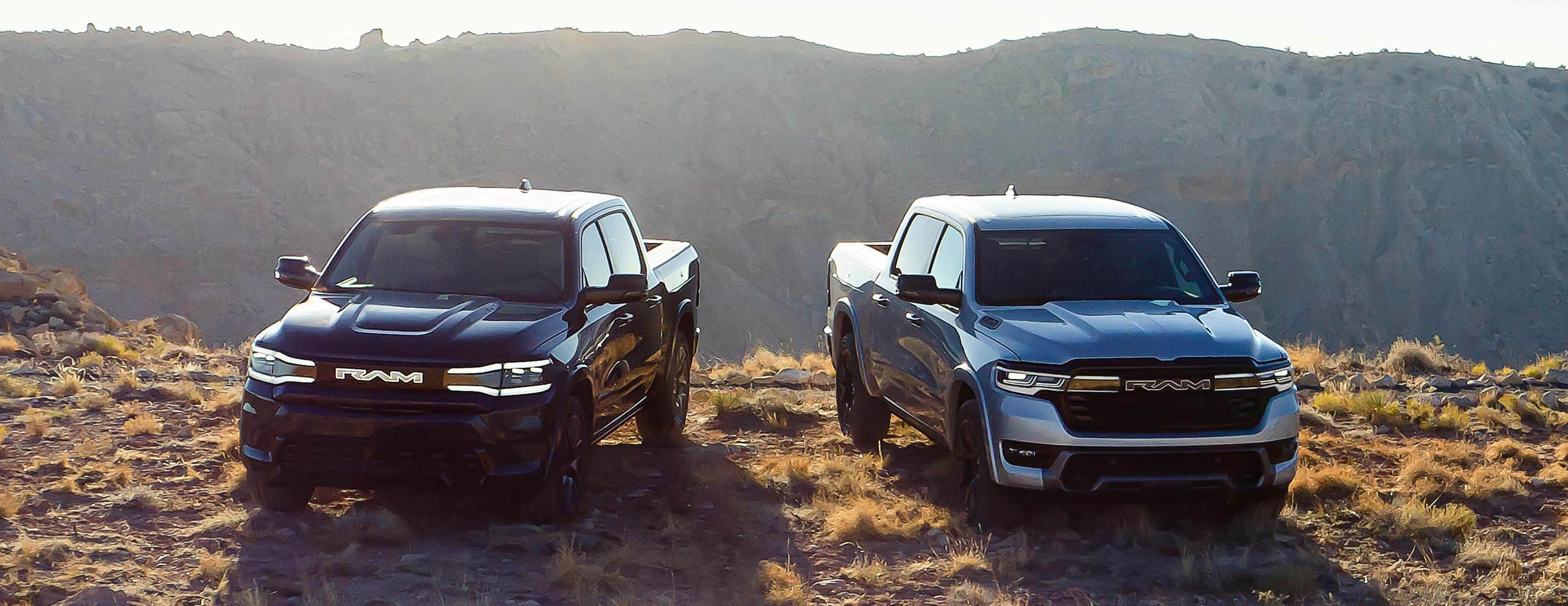 Dos vehículos eléctricos de la marca Ram estacionados uno junto al otro en una colina de césped al amanecer con las montañas a la distancia. A la izquierda, una Ram 1500 Rev Tungsten 4x4 Crew Cab 2025 negra y a la derecha una Ram 1500 Ramcharger Limited 4x4 Crew Cab 2025 azul.