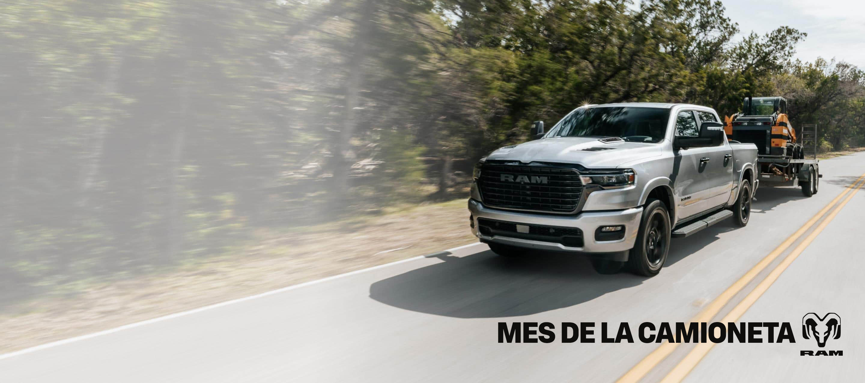 Una Ram 1500 Laramie 4x4 Crew Cab 2026 plateada circulando por una carretera, remolcando una excavadora sobre una minicargadora. Mes de las camionetas Ram.