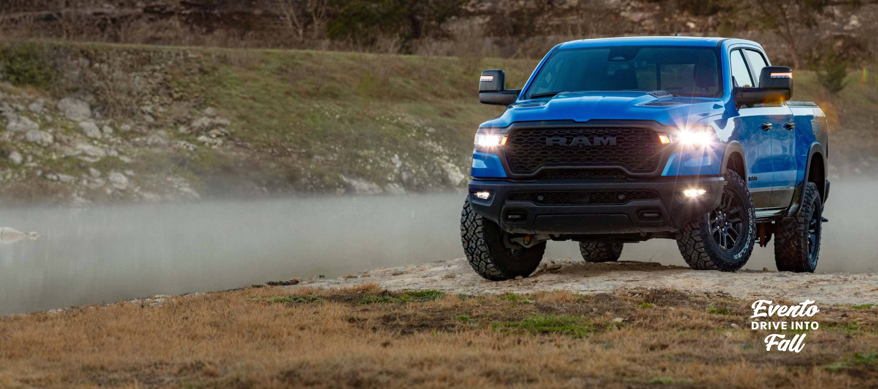 Una Ram 1500 Rebel 4x4 Crew Cab 2025 azul, con sus faros delanteros y antiniebla encendidos, sube una pequeña colina fuera de la carretera. Evento de ventas Drive Into Fall.