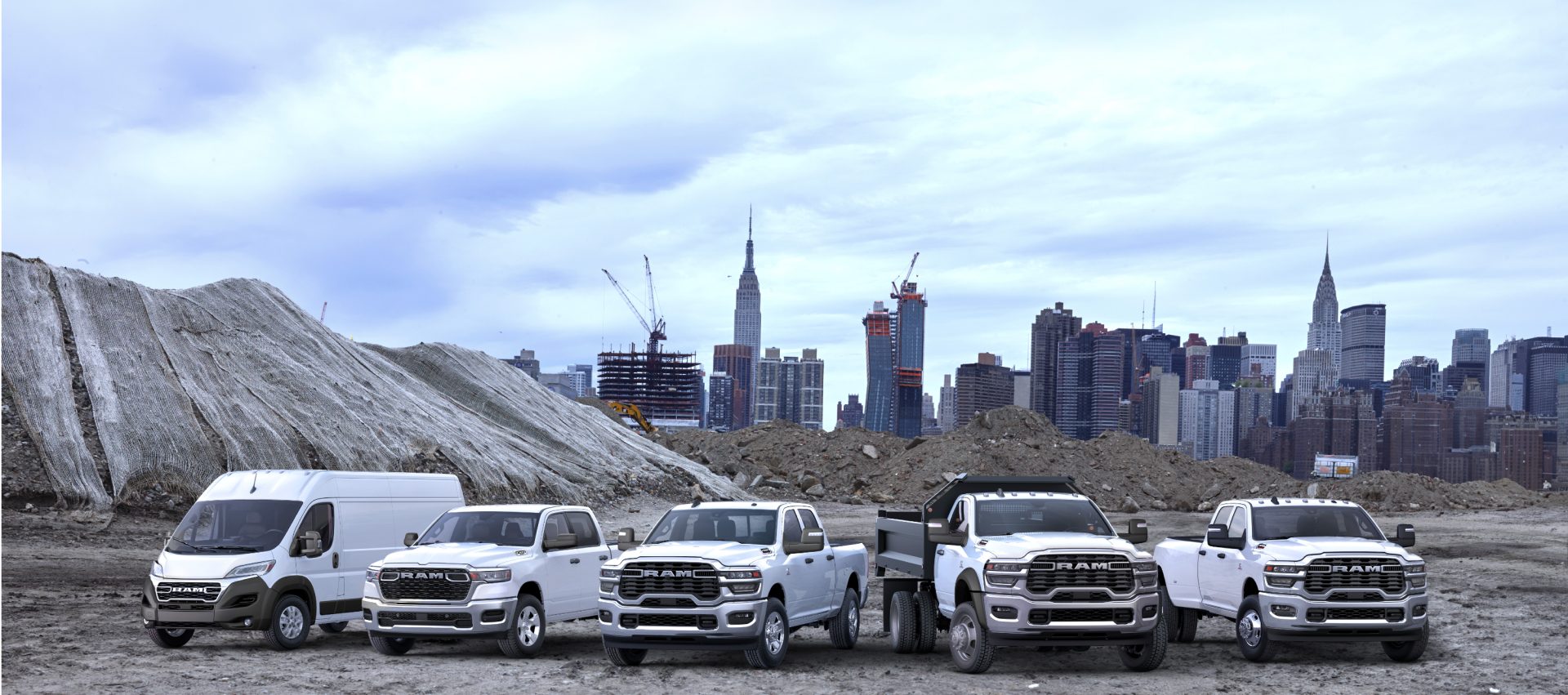 Línea de la marca Ram 2025 toda en blanco, con los vehículos estacionados uno al lado del otro en una obra de construcción, con un paisaje urbano de fondo. De izquierda a derecha: una Ram ProMaster 3500 SLT High Roof Cargo Van, una Ram 1500 Tradesman 4x4 Crew Cab, una Ram 2500 Tradesman 4x4 Crew Cab, una Ram 5500 Tradesman Chassis Regular Cab con mejora de carrocería volcadora y una Ram 3500 Tradesman 4x4 Crew Cab.