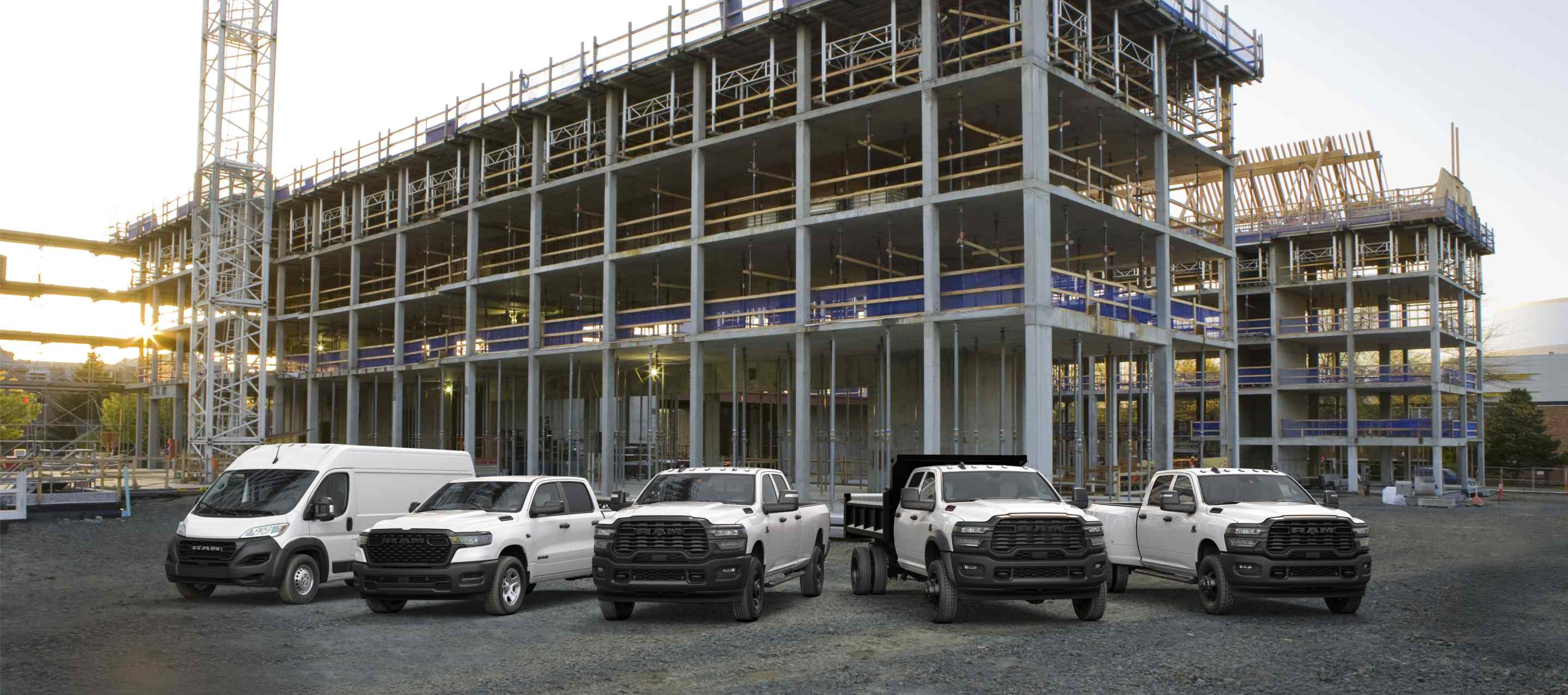 Una formación de camionetas y vans Ram 2026, todas de color blanco, en una obra comercial en construcción. De izquierda a derecha: una Ram ProMaster 3500 Tradesman High Roof Cargo Van, una Ram 1500 Tradesman 4x4 Crew Cab, una Ram 2500 Tradesman 4x4 Crew Cab, una Ram 3500 Tradesman Crew Cab Chassis Cab con una mejora de carrocería volcadora​​​​​​​ y una Ram 3500 Tradesman 4x4 Crew Cab.