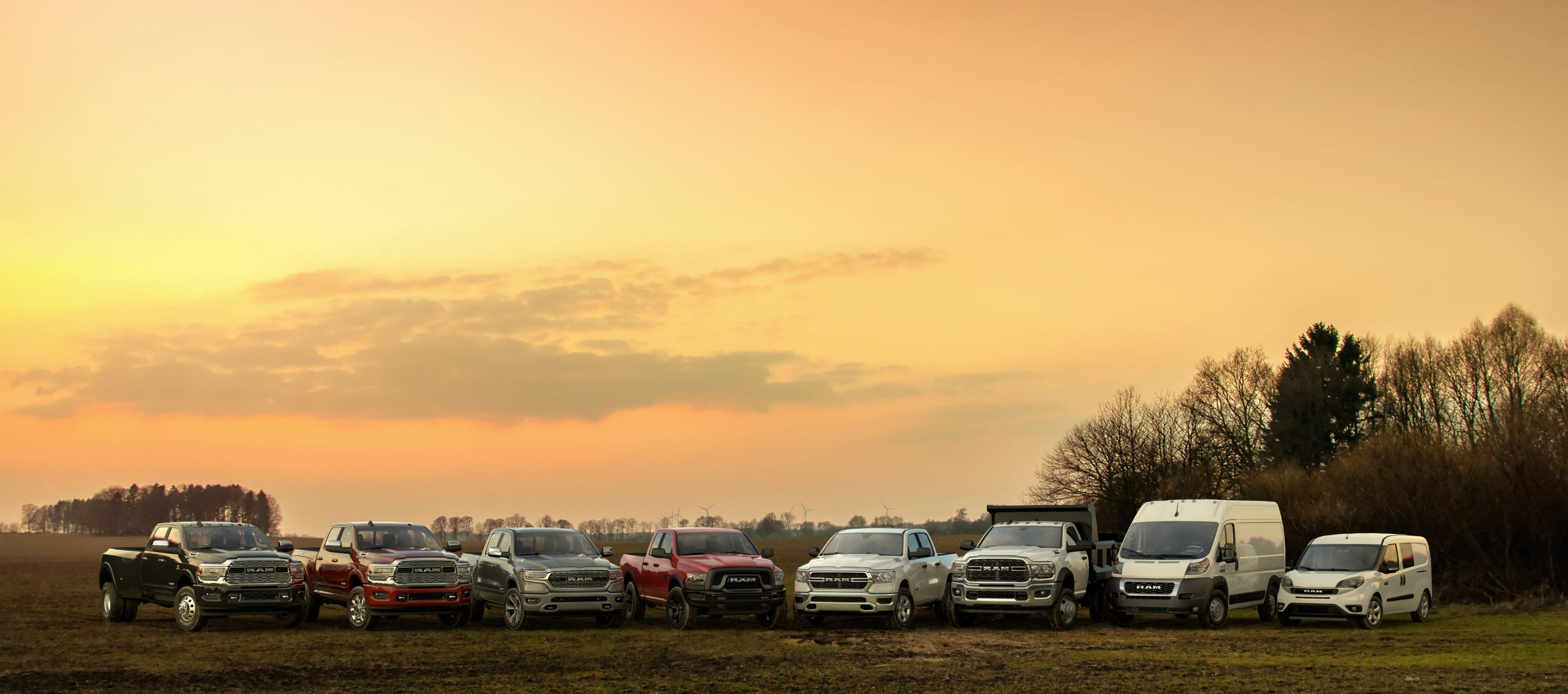 A lineup of eight Ram Brand trucks and vans parked in a clearing at sunset.