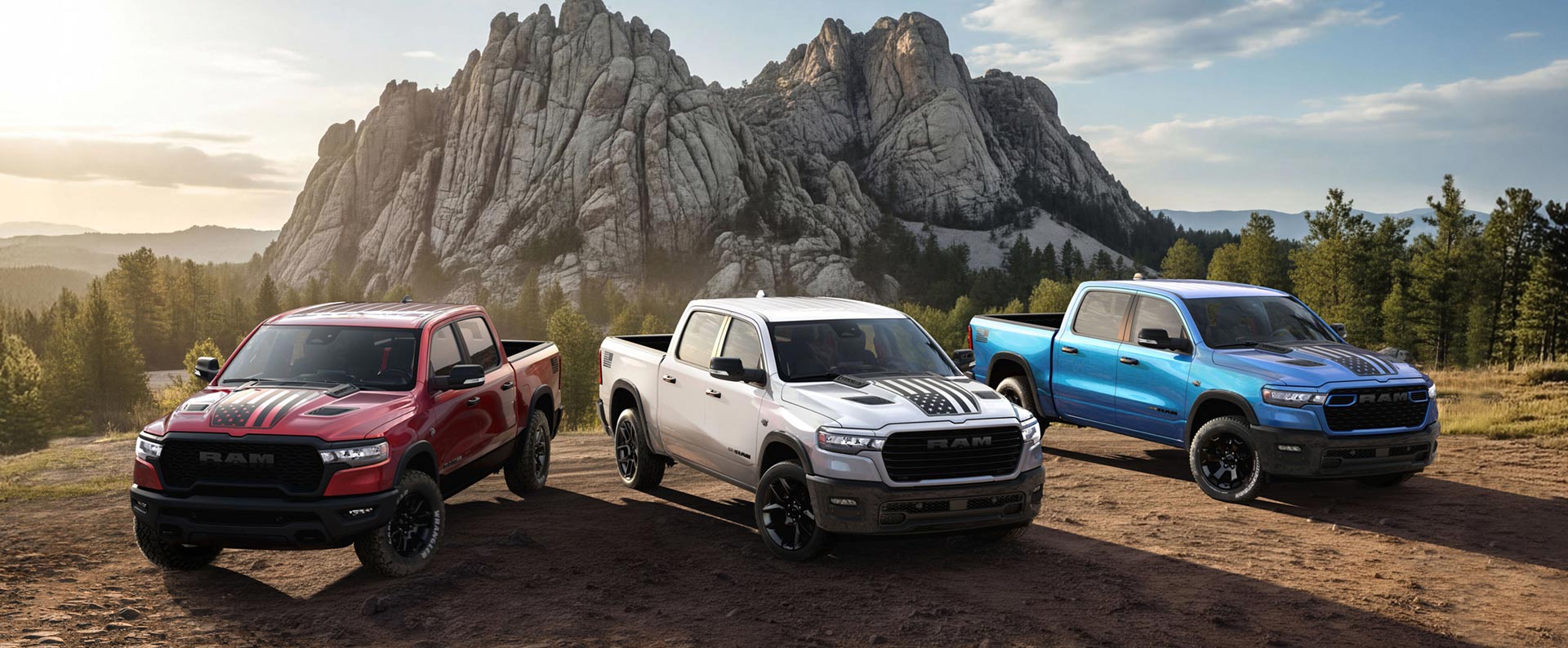 Tres camionetas Ram 1500 America 250th Anniversary Edition 2026 sobre una colina, con montañas al fondo. De izquierda a derecha: una Ram 1500 Rebel 4x4 Crew Cab roja, una Ram 1500 Laramie 4x4 Crew Cab blanca y una Ram 1500 Big Horn 4x4 Crew Cab azul.