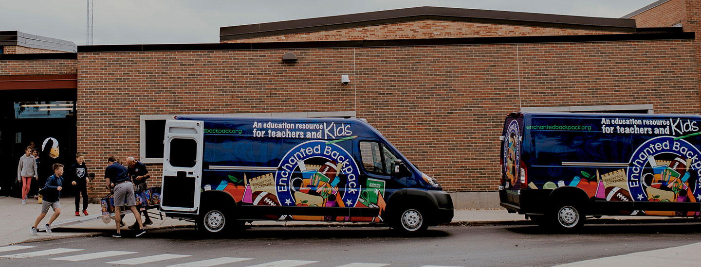 Dos vans Ram ProMaster 2018 con publicidad de Enchanted Backpack estacionadas frente a una escuela.