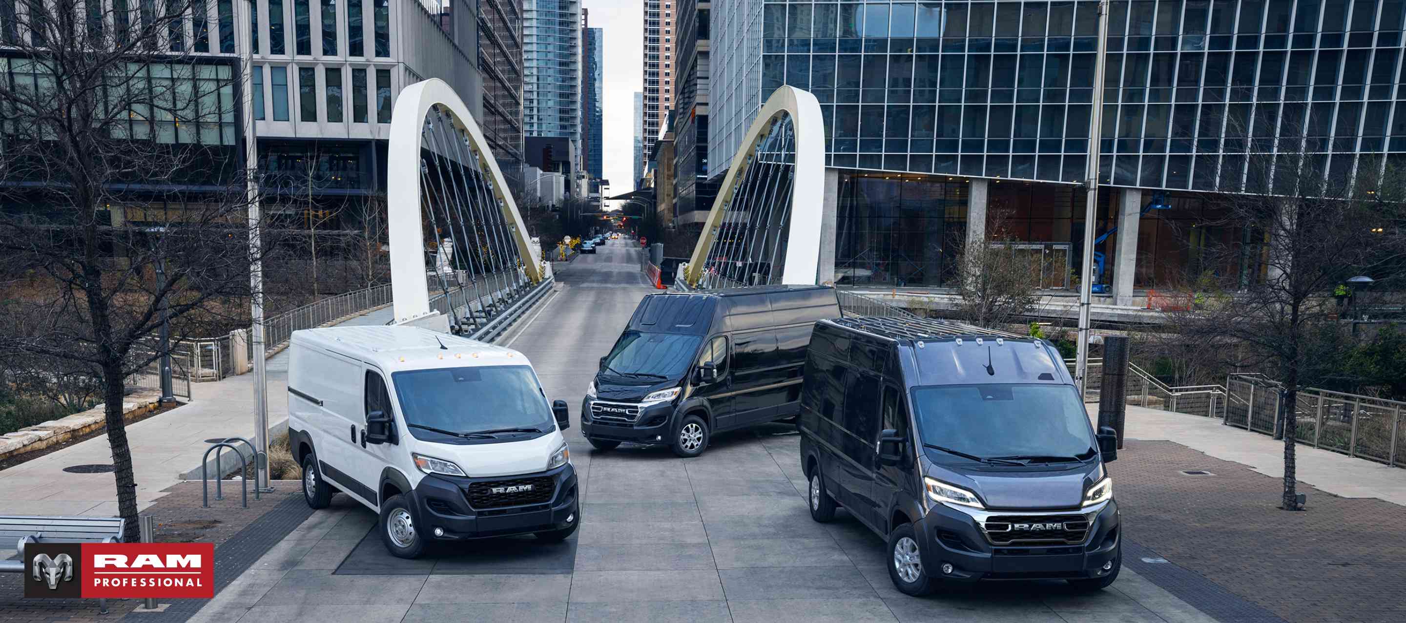 Tres modelos Ram ProMaster 2024, de izquierda a derecha: una Ram ProMaster 1500 Tradesman Standard Roof Cargo Van blanca, una Ram ProMaster 3500 SLT+ Super High Roof Cargo Van negra y una Ram ProMaster 3500 SLT High Roof Cargo Van negra, estacionadas en el centro de una gran ciudad ajetreada, con un puente de fondo. Ram Professional.