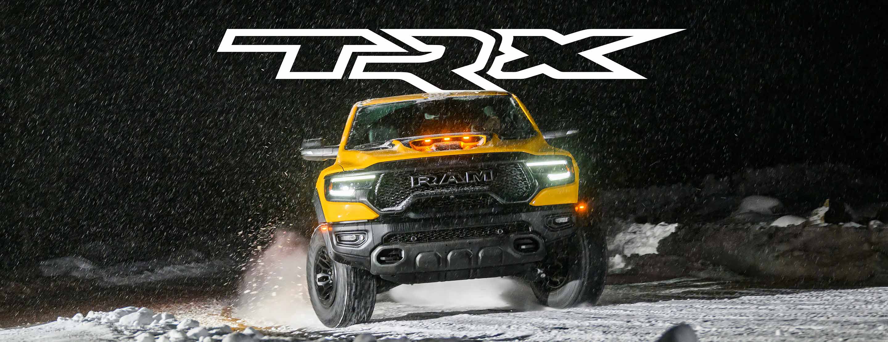 TRX. Ángulo frontal de una Ram 1500 TRX Havoc Edition 2023 amarilla circulando de noche, levantando nieve con los neumáticos.